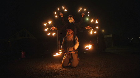 Två personer uppträder med eld i mörkret, en står på knä och en bär mask och håller eldredskap.