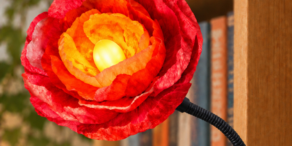 En blomformad lampa med klämfäste sitter fast på en bokhylla, omgiven av böcker och växter.