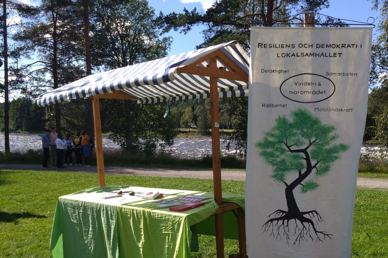 Ett marknadsstånd med grön duk och informationsmaterial står utomhus på gräs, med en affisch om resiliens och demokrati.