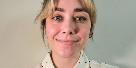 Kvinna med blont hår och prickig vit skjorta ler mot kameran framför en ljus bakgrund.