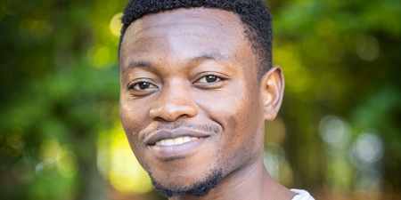 En ung man med kort hår och ljusgrå tröja står utomhus framför gröna träd och ler mot kameran.