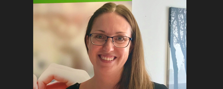 Leende kvinna med glasögon och långt hår står inomhus framför en affisch och en tavla.