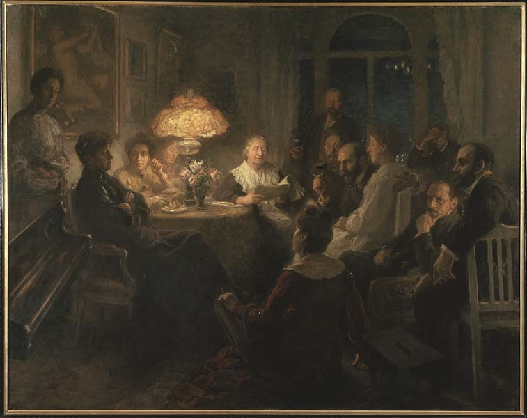 En grupp människor sitter samlade runt ett bord i ett svagt upplyst rum, några lyssnar på en kvinna som läser högt.