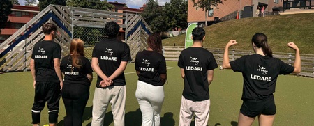 Sex personer står med ryggen mot kameran på en gräsplan, alla bär svarta tröjor med texten 'LEDARE'.