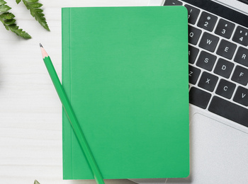 Grön anteckningsbok och penna på en bärbar dator, med växt och ormbunksblad på ett vitt skrivbord.