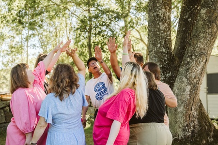 En grupp människor står i en ring utomhus och sträcker upp händerna mot mitten under ett träd.