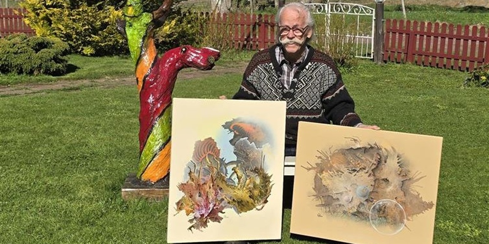 Person sitter utomhus och visar två konstverk, med en färgglad träskulptur bredvid.