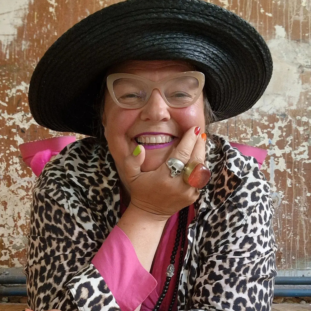 Leende person med stor svart hatt, glasögon och leopardmönstrad jacka mot sliten vägg.