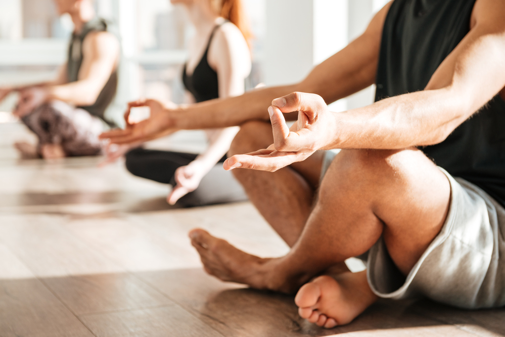 Personer sitter i lotusställning och mediterar på ett ljust trägolv i en yogastudio.