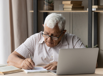 Äldre man med glasögon skriver i anteckningsbok vid laptop, omgiven av böcker på ett skrivbord.