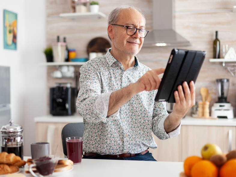 Äldre man sitter vid köksbordet, ler och använder en surfplatta. Frukost med kaffe och frukt på bordet.