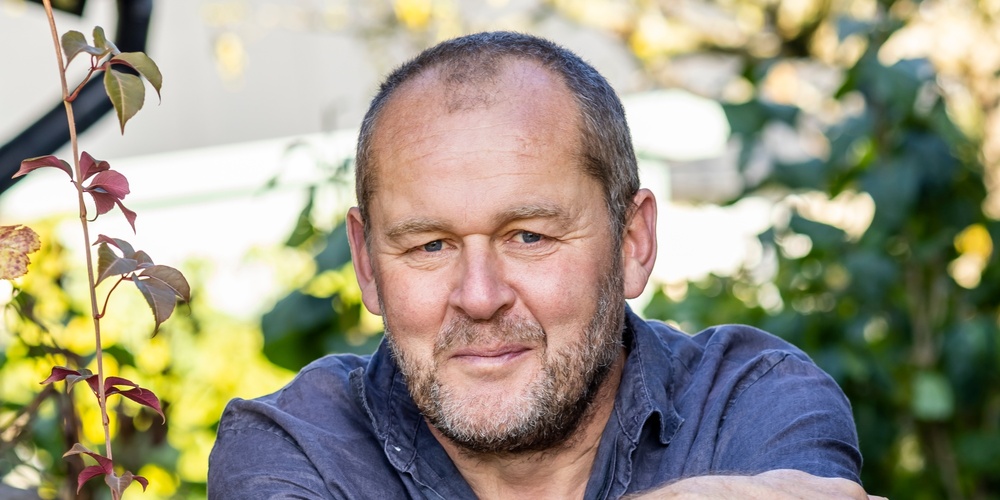 En person står i en trädgård med armarna vilande på ett trädgårdsredskap, omgiven av gröna växter.