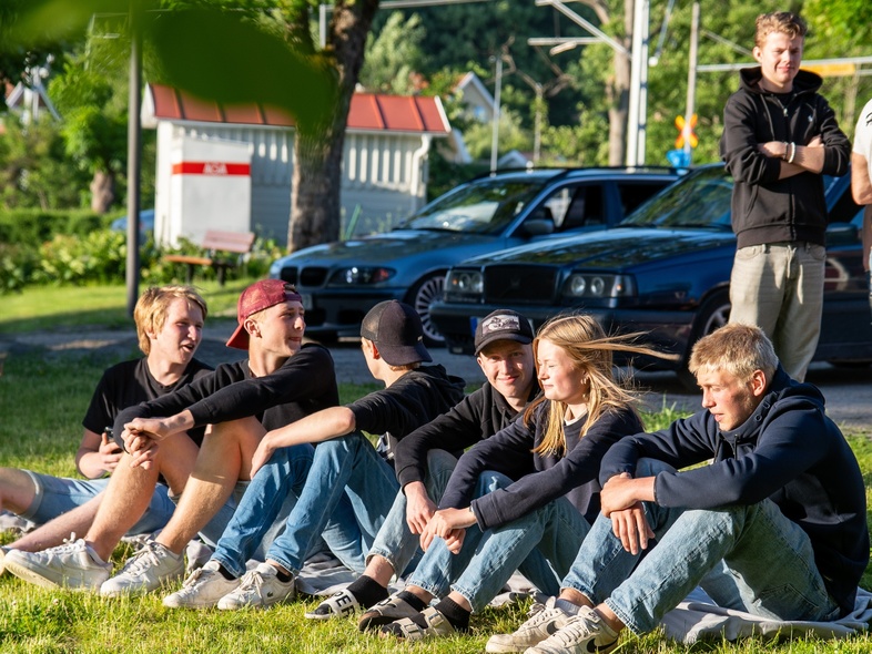 En grupp ungdomar sitter på gräset i solen, med bilar och träd i bakgrunden.