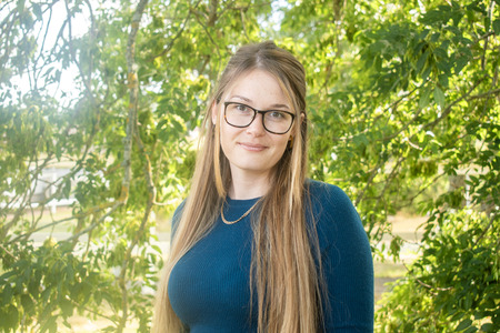Kvinna med långt blont hår och glasögon står utomhus framför gröna träd, bär blå tröja och halskedja.