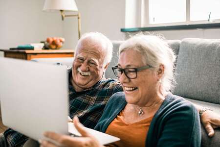 Två personer sitter på golvet framför en grå soffa och tittar skrattandes på en laptop.