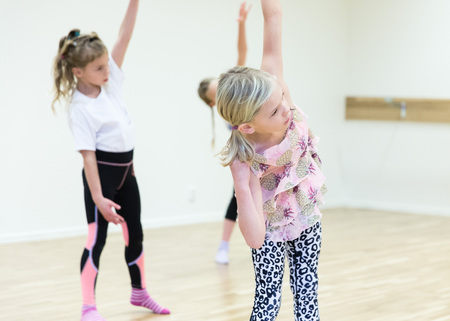 Tre barn gör stretchövningar i ett ljust dansrum med trägolv.