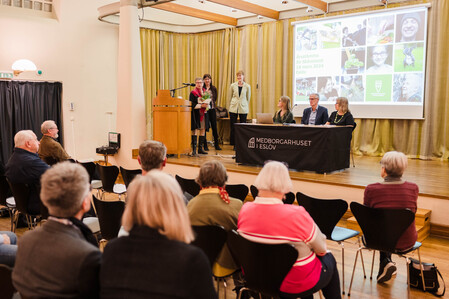 En konferens eller presentation pågår i en sal med publik, fyra personer sitter vid ett bord på scenen och två står vid ett podium.