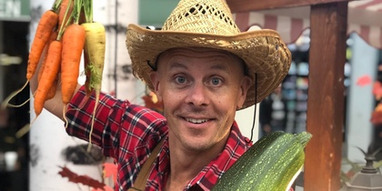 En person i cowboyhatt och rutigt skjorta håller stora morötter och en jättestor zucchini på en marknad.