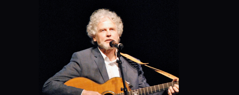 Man med grått lockigt hår spelar akustisk gitarr och sjunger i mikrofon på scen.