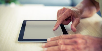 En äldre person använder en surfplatta på ett ljust träbord.