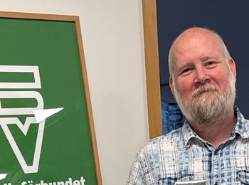 Man står leende med ett paket i handen bredvid en skylt för Studieförbundet Vuxenskolan.