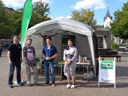 Fyra personer står framför ett tält med informationsbord på ett torg, med en kyrka i bakgrunden.