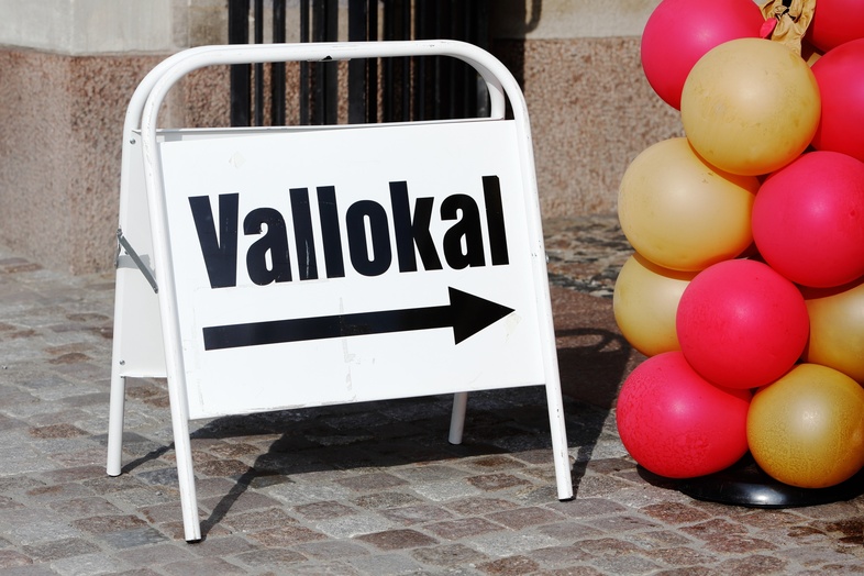 Skylt med texten 'Vallokal' och en pil åt höger, bredvid röda och gula ballonger på stenlagd mark.