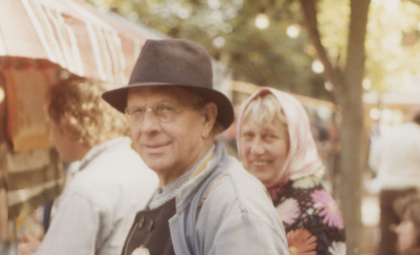 Två personer på en utomhusmarknad, en med hatt och en med sjal, suddiga ansikten, träd och marknadsstånd i bakgrunden.