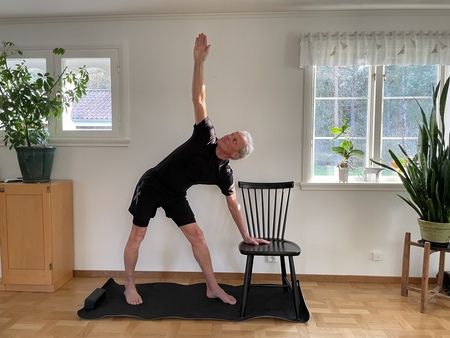 Äldre man gör en yogaposition med stöd av en stol i ett ljust vardagsrum med växter.