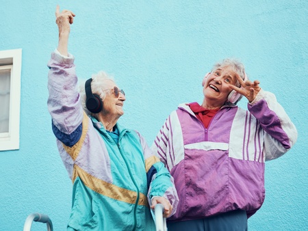Två glada äldre kvinnor i färgglada träningsjackor poserar och dansar framför en blå vägg.