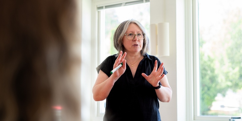 Kvinna med glasögon står och pratar framför ett fönster i ett ljust rum.