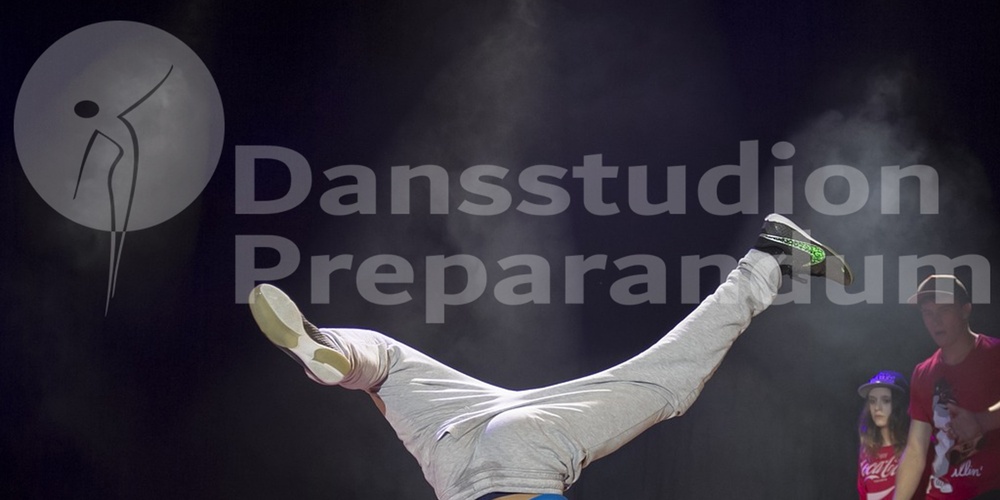 En person gör en breakdance-move på scen medan andra tittar på i bakgrunden.