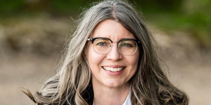 Leende kvinna med långt grått hår och glasögon utomhus, klädd i vit skjorta och mörk kavaj.