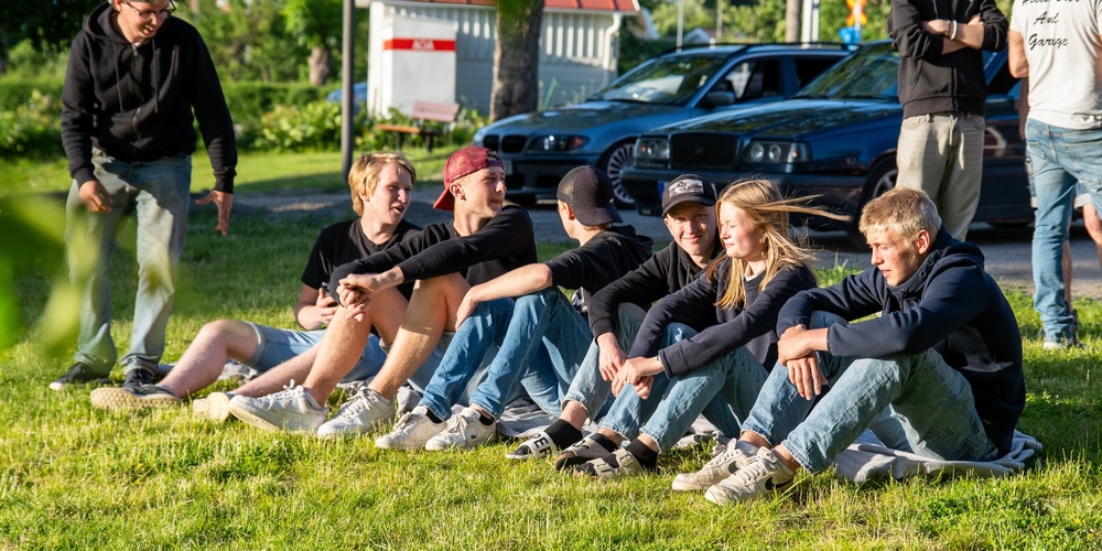 En grupp ungdomar sitter på gräset i solen, några vuxna står i bakgrunden vid bilar.