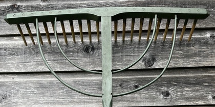 En gammaldags träräfsa med grönt huvud och trähandtag står mot en vägg av grått trä.