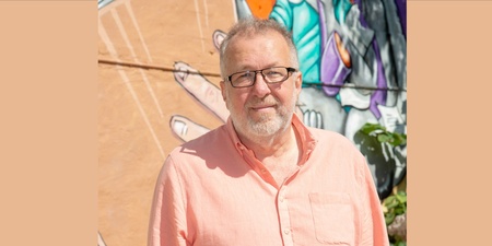 Person i ljusrosa skjorta står framför en färgglad väggmålning utomhus.