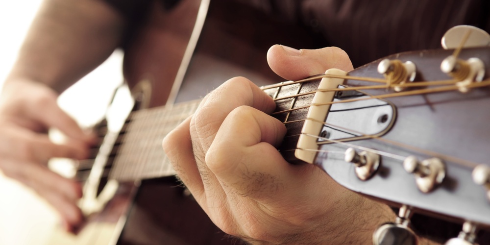 Närbild av en person som spelar akustisk gitarr, fokus på vänster hand på gitarrens greppbräda.
