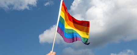 En hand håller upp en regnbågsflagga mot en blå himmel med vita moln.