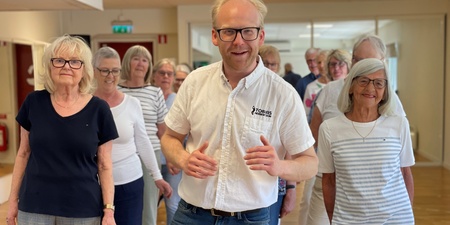 En grupp äldre personer och en yngre man står tillsammans inomhus, leende och avslappnade.