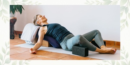 Person utför en avslappnande yogaposition med stöd av kuddar och block på en yogamatta inomhus.
