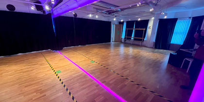 Ett tomt dans- eller träningsrum med trägolv, svarta gardiner och lila belysning.