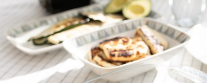 Ett dukat bord med grillad majs, sparris, kyckling, avokado och en vas med lila blommor i solljus.