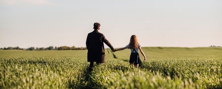 Två personer, en vuxen och ett barn, går hand i hand genom en grön åker under blå himmel.