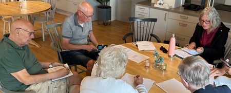 Sex äldre personer sitter runt ett bord i ett ljust kök och skriver på papper.