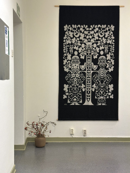 En väggbonad med två stiliserade figurer och ett träd hänger på en vit vägg ovanför ett brunt krus med torkade grenar.