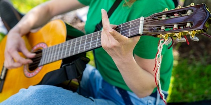 Person i grön tröja spelar akustisk gitarr utomhus, fokus på händer och gitarr.