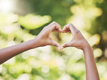 Två händer formar ett hjärta mot en suddig, grön bakgrund utomhus.