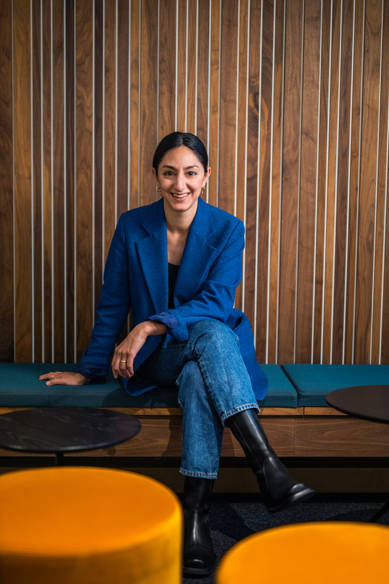 Kvinna i blå kavaj sitter leende på en bänk mot en trävägg med färgglada pallar framför sig.