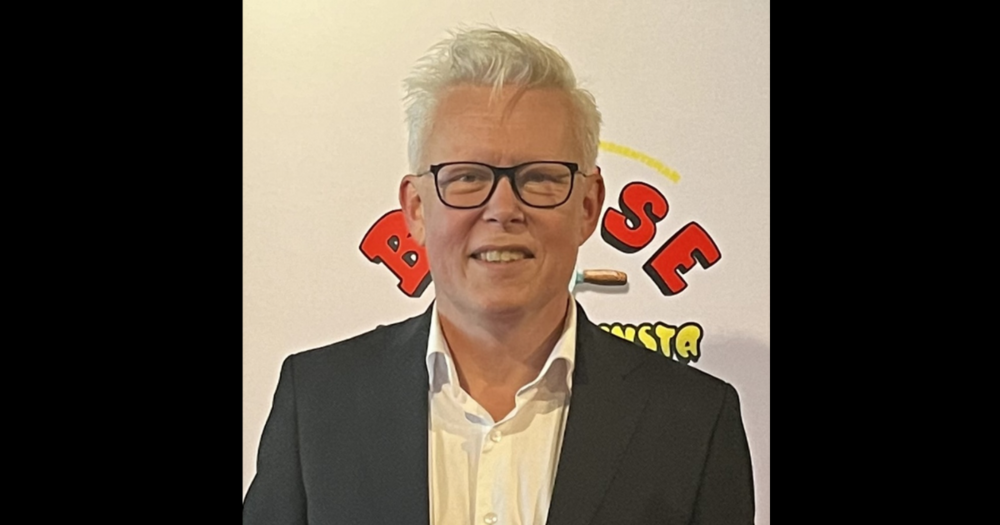 Man med vitt hår och glasögon i kostym står framför en ljus bakgrund med färgglad text.