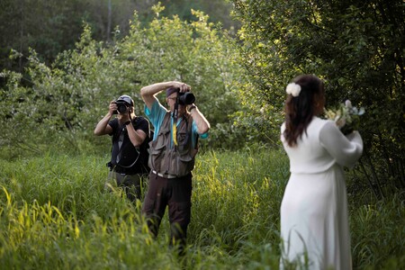 Två fotografer tar bilder av en kvinna i vit klänning som står i en grönskande naturmiljö.